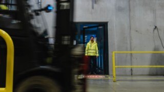Un hombre con ropa de seguridad sale por una puerta para entrar en una nave industrial. Pasa una carretilla elevadora. Un hombre con ropa de seguridad sale por una puerta para entrar en una nave industrial. Pasa una carretilla elevadora.