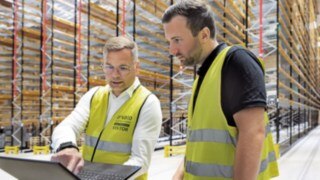 Dos hombres con chalecos reflectantes amarillos están de pie en un almacén de estanterías altas mirando un ordenador portátil. 