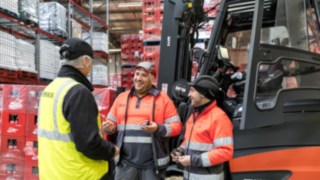 El confort y la seguridad de los vehículos Linde aumentan la satisfacción laboral y la motivación de los conductores. Tres personas hablan delante de una carretilla elevadora.